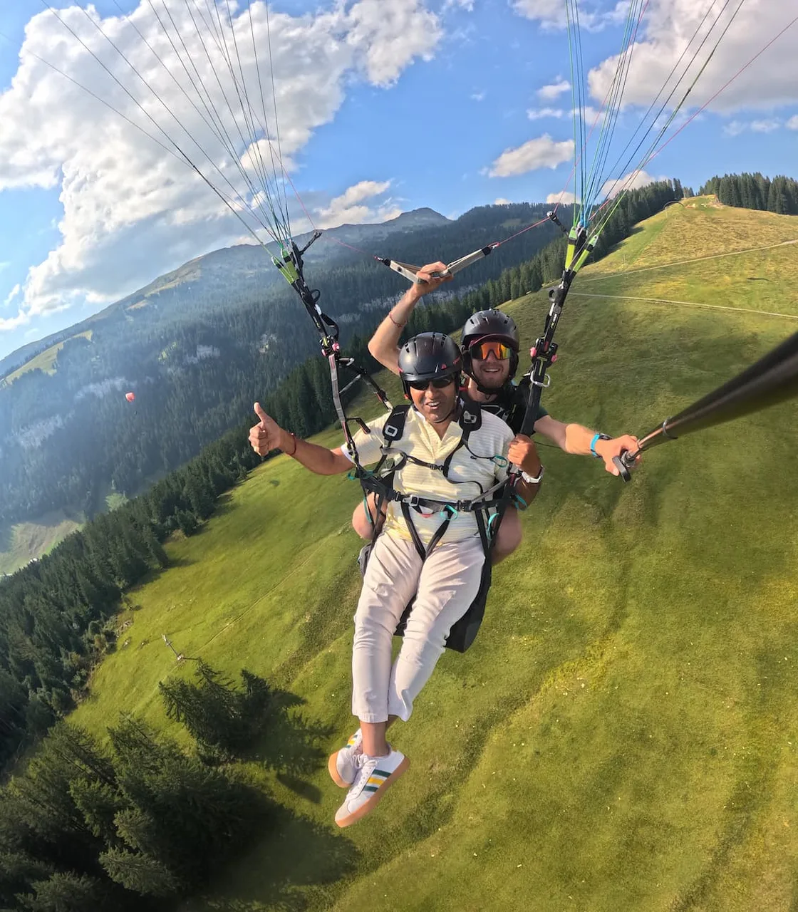 Passagère souriante en vol biplace avec son pilote au-dessus d’une pente verte du Valais.